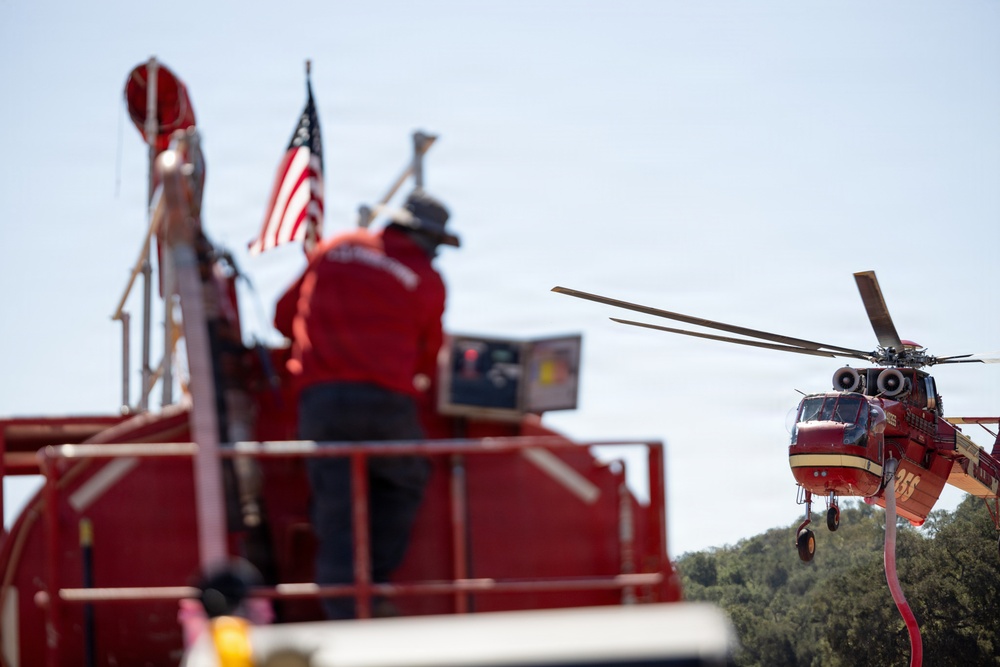 A firefighting operation is underway, showing a person in red standing on top of a red fire truck with an American flag visible. In the background, a red firefighting helicopter (numbered 355) is hovering nearby, with a hose or snorkel attachment visible beneath it. The scene appears to be an aerial water-drop coordination effort in a wildland or wildland-urban interface firefighting context.