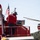 A firefighting operation is underway, showing a person in red standing on top of a red fire truck with an American flag visible. In the background, a red firefighting helicopter (numbered 355) is hovering nearby, with a hose or snorkel attachment visible beneath it. The scene appears to be an aerial water-drop coordination effort in a wildland or wildland-urban interface firefighting context.