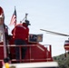 A firefighting operation is underway, showing a person in red standing on top of a red fire truck with an American flag visible. In the background, a red firefighting helicopter (numbered 355) is hovering nearby, with a hose or snorkel attachment visible beneath it. The scene appears to be an aerial water-drop coordination effort in a wildland or wildland-urban interface firefighting context.