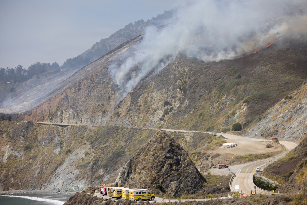 A wildfire is actively burning on a steep coastal hillside, with visible flames and heavy smoke rising from multiple points along the slope. Emergency response vehicles, including yellow fire trucks, are staged along a coastal highway at the base of the hill. The scene appears to be along a rugged Pacific Coast highway, likely in California, with the ocean visible at lower left.