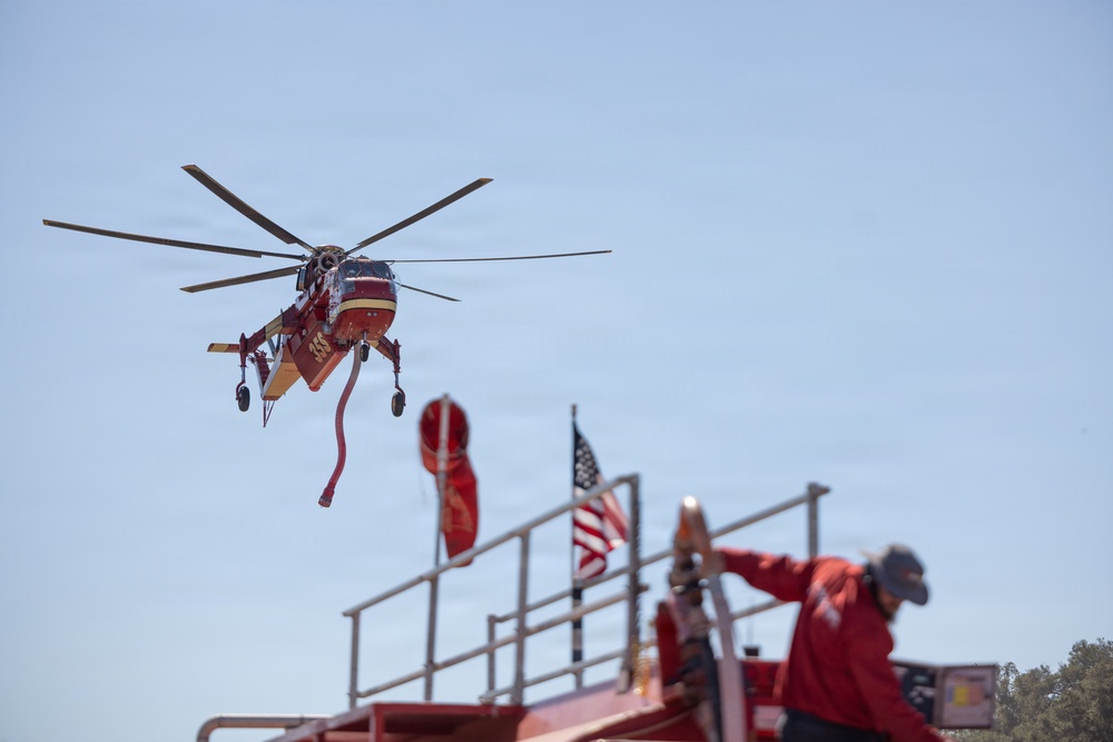 A red firefighting helicopter, appearing to be a Sikorsky S-64 Aircrane (Erickson Air-Crane), hovers in the sky with a water/retardant snorkel hose dangling beneath it. In the foreground, a firefighter in red gear stands atop a red fire apparatus, with an American flag visible nearby. The scene suggests an active wildfire suppression operation.