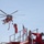 A red firefighting helicopter, appearing to be a Sikorsky S-64 Aircrane (Erickson Air-Crane), hovers in the sky with a water/retardant snorkel hose dangling beneath it. In the foreground, a firefighter in red gear stands atop a red fire apparatus, with an American flag visible nearby. The scene suggests an active wildfire suppression operation.