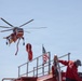 A red firefighting helicopter, appearing to be a Sikorsky S-64 Aircrane (Erickson Air-Crane), hovers in the sky with a water/retardant snorkel hose dangling beneath it. In the foreground, a firefighter in red gear stands atop a red fire apparatus, with an American flag visible nearby. The scene suggests an active wildfire suppression operation.