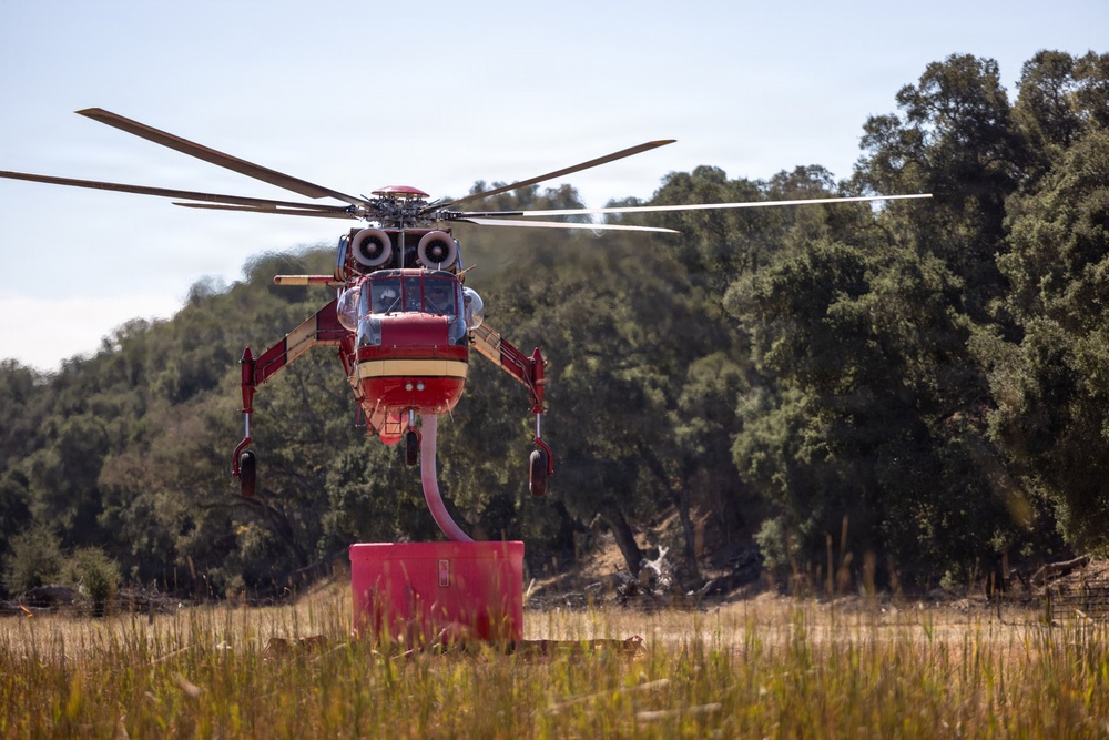 A large red and cream firefighting helicopter is hovering low over a dry grassy field, actively drawing water through a snorkel/hose from a red portable water tank on the ground. The helicopter appears to be a heavy-lift type, likely a Sikorsky S-64 Aircrane or similar model, equipped with a belly-mounted water tank system. The setting suggests a wildfire suppression operation in a rural or semi-rural area with dry vegetation and oak trees in the background.