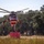 A large red and cream firefighting helicopter is hovering low over a dry grassy field, actively drawing water through a snorkel/hose from a red portable water tank on the ground. The helicopter appears to be a heavy-lift type, likely a Sikorsky S-64 Aircrane or similar model, equipped with a belly-mounted water tank system. The setting suggests a wildfire suppression operation in a rural or semi-rural area with dry vegetation and oak trees in the background.