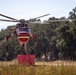 A large red and cream firefighting helicopter is hovering low over a dry grassy field, actively drawing water through a snorkel/hose from a red portable water tank on the ground. The helicopter appears to be a heavy-lift type, likely a Sikorsky S-64 Aircrane or similar model, equipped with a belly-mounted water tank system. The setting suggests a wildfire suppression operation in a rural or semi-rural area with dry vegetation and oak trees in the background.