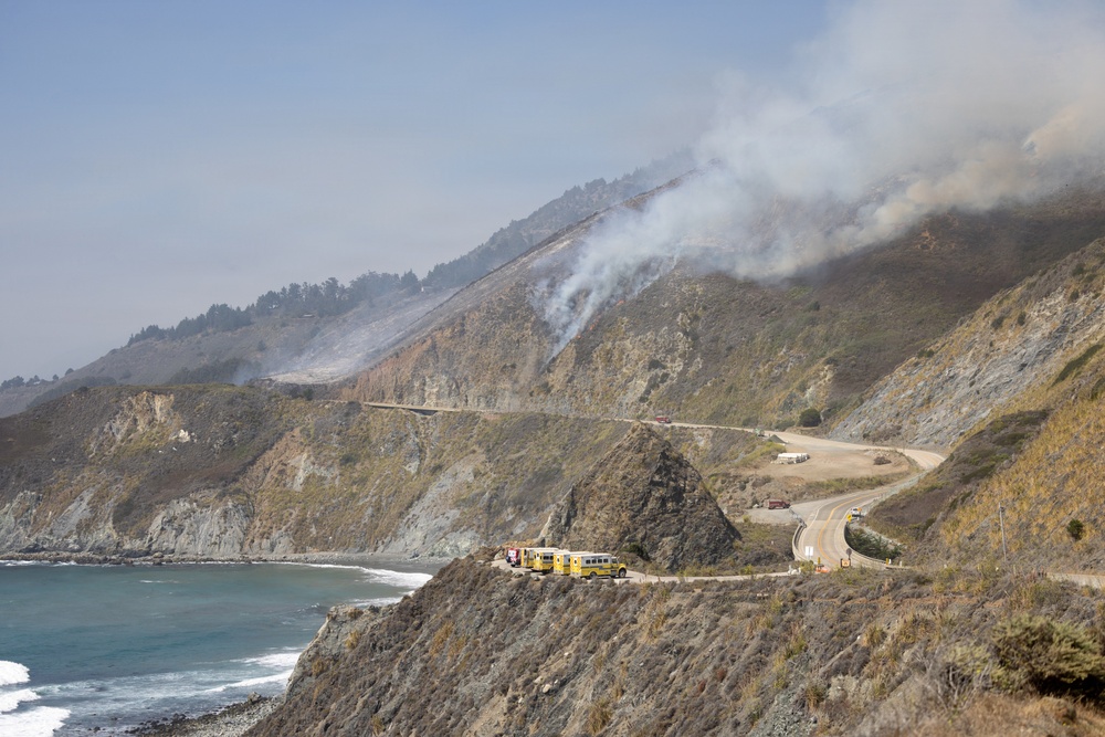 A wildfire burns on a steep coastal hillside, with smoke rising visibly from multiple points on the mountain slope. Emergency response vehicles, including yellow fire trucks, are staged on a narrow coastal highway that winds along dramatic sea cliffs above the Pacific Ocean. The scene depicts an active firefighting operation along what appears to be a rugged California coastline.