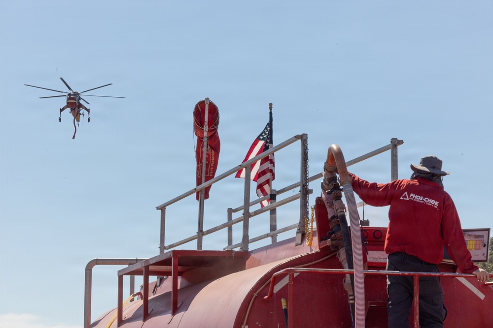 A worker wearing a Phos-Chek branded red uniform stands atop a large red fire retardant tanker truck, managing hoses. An American flag is mounted on the vehicle alongside a red wind sock and fire retardant branding banner. A large firefighting helicopter, consistent with a Sikorsky Skycrane type, is visible in the background sky.