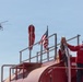 A worker wearing a Phos-Chek branded red uniform stands atop a large red fire retardant tanker truck, managing hoses. An American flag is mounted on the vehicle alongside a red wind sock and fire retardant branding banner. A large firefighting helicopter, consistent with a Sikorsky Skycrane type, is visible in the background sky.