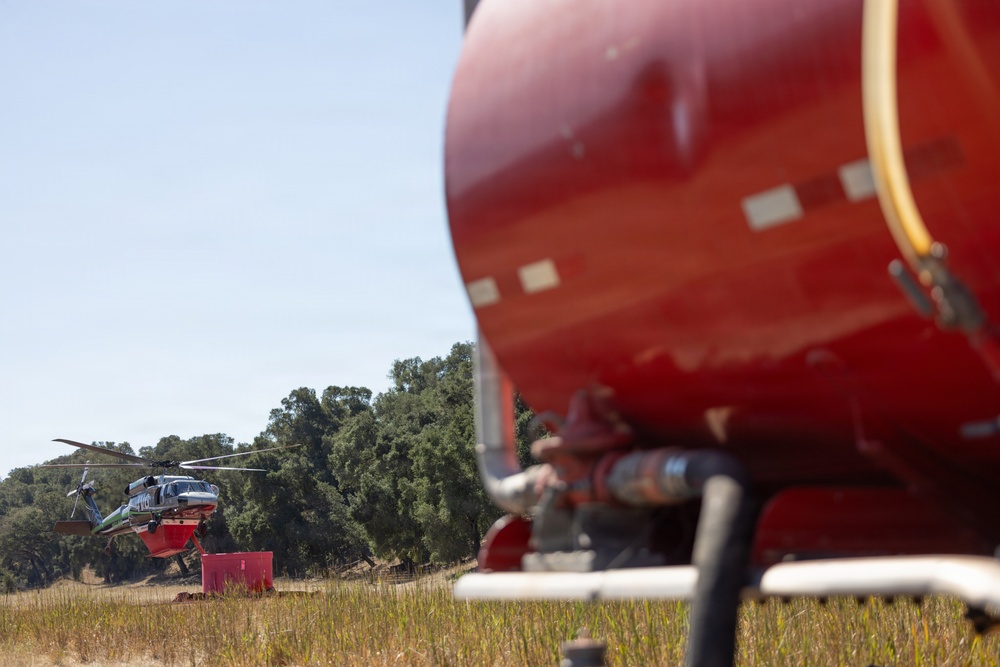 A firefighting helicopter is visible in the background of an open field, appearing to be near a portable water tank or dip tank on the ground. In the foreground, a large red water/retardant tank (likely mounted on a vehicle or trailer) is prominently visible but out of focus. The scene suggests a wildfire aerial operations staging area, with the helicopter equipped with a water bucket.