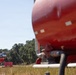 A firefighting helicopter is visible in the background of an open field, appearing to be near a portable water tank or dip tank on the ground. In the foreground, a large red water/retardant tank (likely mounted on a vehicle or trailer) is prominently visible but out of focus. The scene suggests a wildfire aerial operations staging area, with the helicopter equipped with a water bucket.