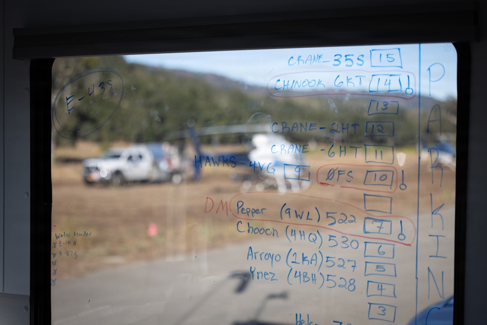 A glass window or whiteboard at what appears to be a wildfire or emergency incident command post displays handwritten operational assignments, unit identifiers, and numbered slots. The writing lists aircraft (Crane, Chinook) and ground units (Hawks, DMC, Pepper, Chooon, Arroyo, Ynez) with radio frequencies and position numbers. Through the glass, a field staging area is visible with white vehicles and what appears to be a helicopter in the background.
