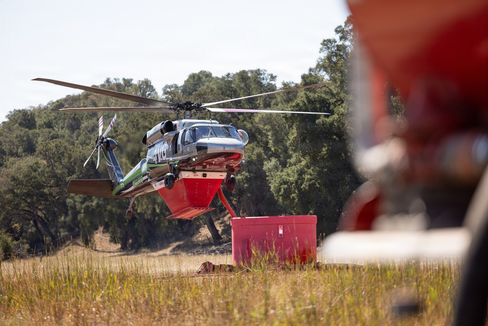 A firefighting helicopter, marked with green and white livery and the number '4VG', is hovering low over a dry grassy field while connected to a red water tank or dip tank on the ground. The helicopter appears to be a Sikorsky S-76 or similar medium-lift aircraft equipped with a belly-mounted water tank for aerial firefighting operations. The scene suggests an active wildfire suppression operation or a refilling/staging operation in a rural, oak-woodland environment.