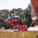 A firefighting helicopter, marked with green and white livery and the number '4VG', is hovering low over a dry grassy field while connected to a red water tank or dip tank on the ground. The helicopter appears to be a Sikorsky S-76 or similar medium-lift aircraft equipped with a belly-mounted water tank for aerial firefighting operations. The scene suggests an active wildfire suppression operation or a refilling/staging operation in a rural, oak-woodland environment.