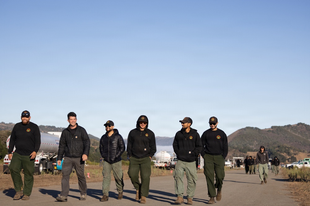 A group of six personnel wearing matching black uniforms with circular patches walk together along a paved road in what appears to be a staging or base camp area. The setting features rolling hills, a clear blue sky, and large tanker trucks visible in the background, suggesting a wildfire or emergency response operation. The group appears relaxed and conversational, likely returning from or heading to an assignment.