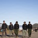 A group of six personnel wearing matching black uniforms with circular patches walk together along a paved road in what appears to be a staging or base camp area. The setting features rolling hills, a clear blue sky, and large tanker trucks visible in the background, suggesting a wildfire or emergency response operation. The group appears relaxed and conversational, likely returning from or heading to an assignment.