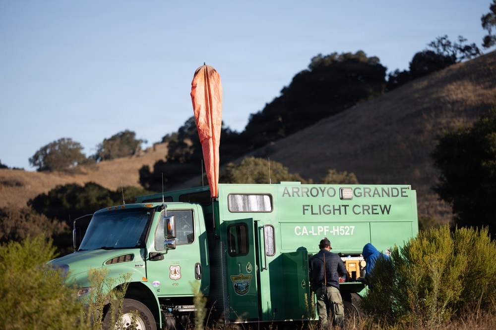 A green US Forest Service truck belonging to the Arroyo Grande Flight Crew is parked in a rural, hilly landscape. The vehicle, identified as CA-LPF-H527, has an orange windsock mounted on top and two personnel working at the rear of the truck. The setting appears to be a staging or operational area in a California wildland environment.