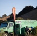 A green US Forest Service truck belonging to the Arroyo Grande Flight Crew is parked in a rural, hilly landscape. The vehicle, identified as CA-LPF-H527, has an orange windsock mounted on top and two personnel working at the rear of the truck. The setting appears to be a staging or operational area in a California wildland environment.