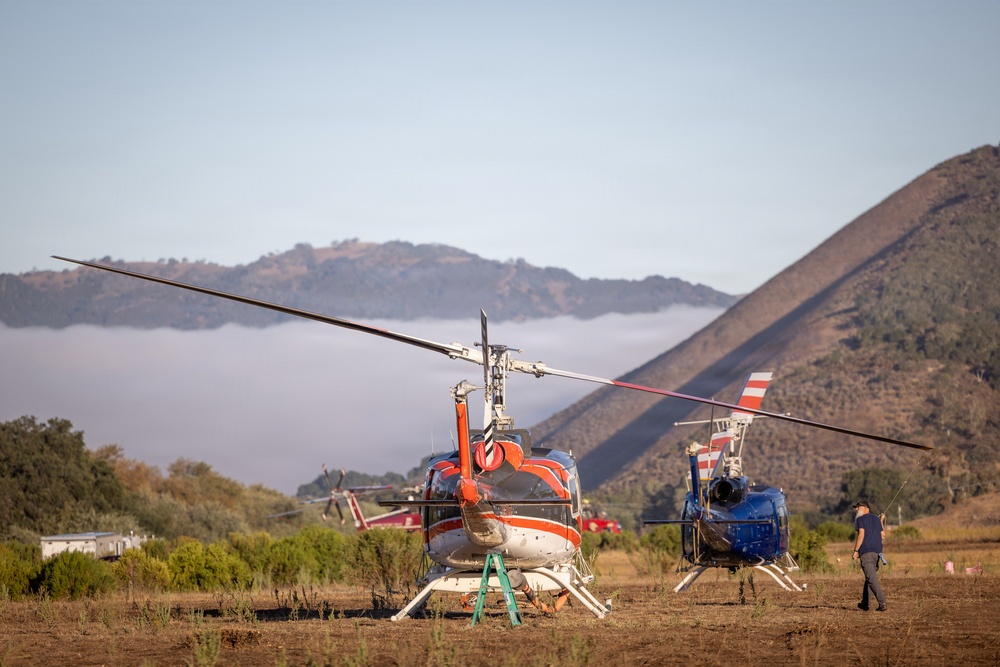 Two helicopters are parked on a dry grass field in a mountainous area, likely serving as a staging or base camp for aerial operations. The foreground helicopter is white and red, while the second is predominantly blue with red and white markings. A person is visible near the second helicopter, and additional aircraft and equipment are visible in the background.