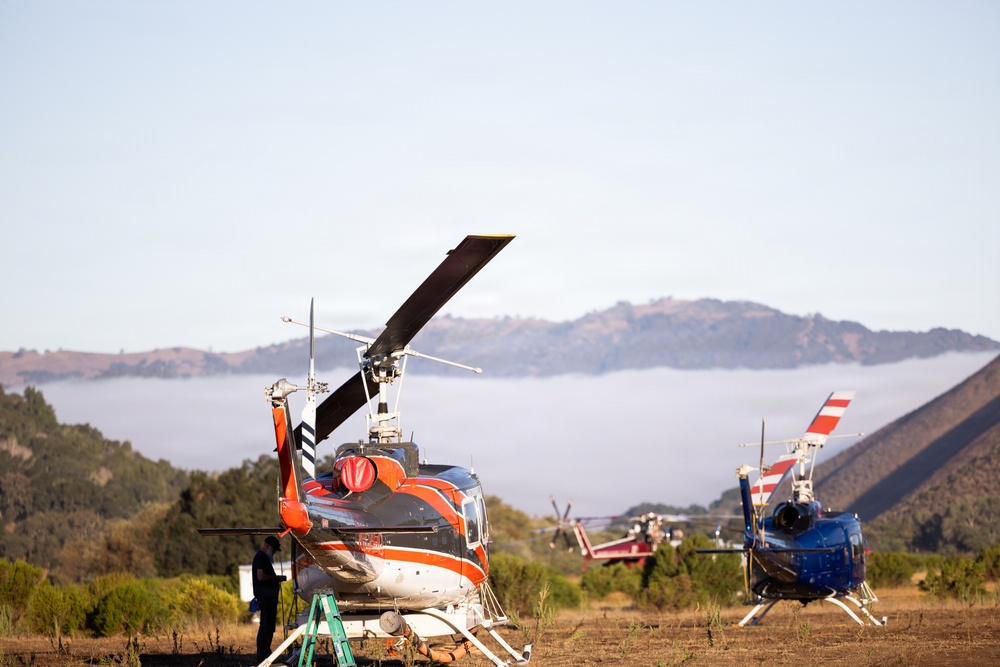 Two helicopters are parked on a dry grassy field in a hilly, semi-arid landscape. The foreground helicopter is white and orange with rotor blades visible, while a blue and red helicopter is parked in the background. A person and a green ladder are visible near the foreground helicopter, suggesting maintenance or inspection activity.