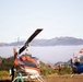 Two helicopters are parked on a dry grassy field in a hilly, semi-arid landscape. The foreground helicopter is white and orange with rotor blades visible, while a blue and red helicopter is parked in the background. A person and a green ladder are visible near the foreground helicopter, suggesting maintenance or inspection activity.