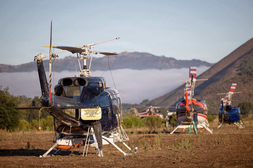 Multiple helicopters are parked on a dry grass field in a mountainous, hilly landscape under a partly cloudy sky. The foreground features a dark blue helicopter with a yellow warning placard and rotor blades tied down, while two red-and-white helicopters are visible in the background. The setting and configuration suggest a temporary staging or base camp area, possibly for firefighting or aerial operations.