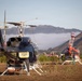 Multiple helicopters are parked on a dry grass field in a mountainous, hilly landscape under a partly cloudy sky. The foreground features a dark blue helicopter with a yellow warning placard and rotor blades tied down, while two red-and-white helicopters are visible in the background. The setting and configuration suggest a temporary staging or base camp area, possibly for firefighting or aerial operations.