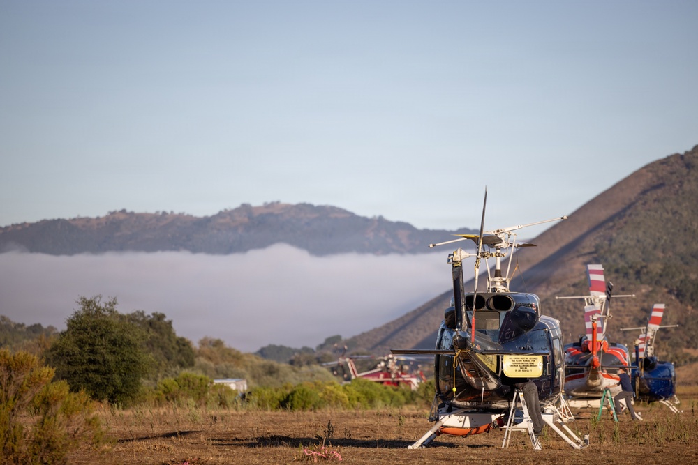 Multiple helicopters are parked on a dry grass field in a mountainous, hilly landscape, likely serving as a staging or base camp area. The foreground features a dark blue helicopter with rotor blades visible, while additional red-and-white helicopters are visible in the background. Low-lying clouds or smoke fill the valley between the hills, suggesting possible wildfire operations or emergency response activity.