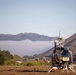 Multiple helicopters are parked on a dry grass field in a mountainous, hilly landscape, likely serving as a staging or base camp area. The foreground features a dark blue helicopter with rotor blades visible, while additional red-and-white helicopters are visible in the background. Low-lying clouds or smoke fill the valley between the hills, suggesting possible wildfire operations or emergency response activity.