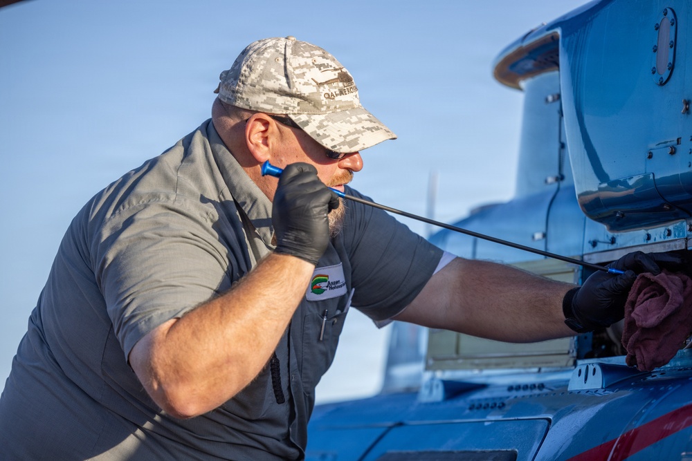 A male aviation mechanic is performing maintenance work on what appears to be a blue aircraft. He is wearing black gloves, a gray shirt, and a camouflage baseball cap, and is closely inspecting or working on a component using a screwdriver-like tool. The setting appears to be outdoors on a clear day at an airfield or maintenance area.