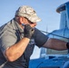 A male aviation mechanic is performing maintenance work on what appears to be a blue aircraft. He is wearing black gloves, a gray shirt, and a camouflage baseball cap, and is closely inspecting or working on a component using a screwdriver-like tool. The setting appears to be outdoors on a clear day at an airfield or maintenance area.