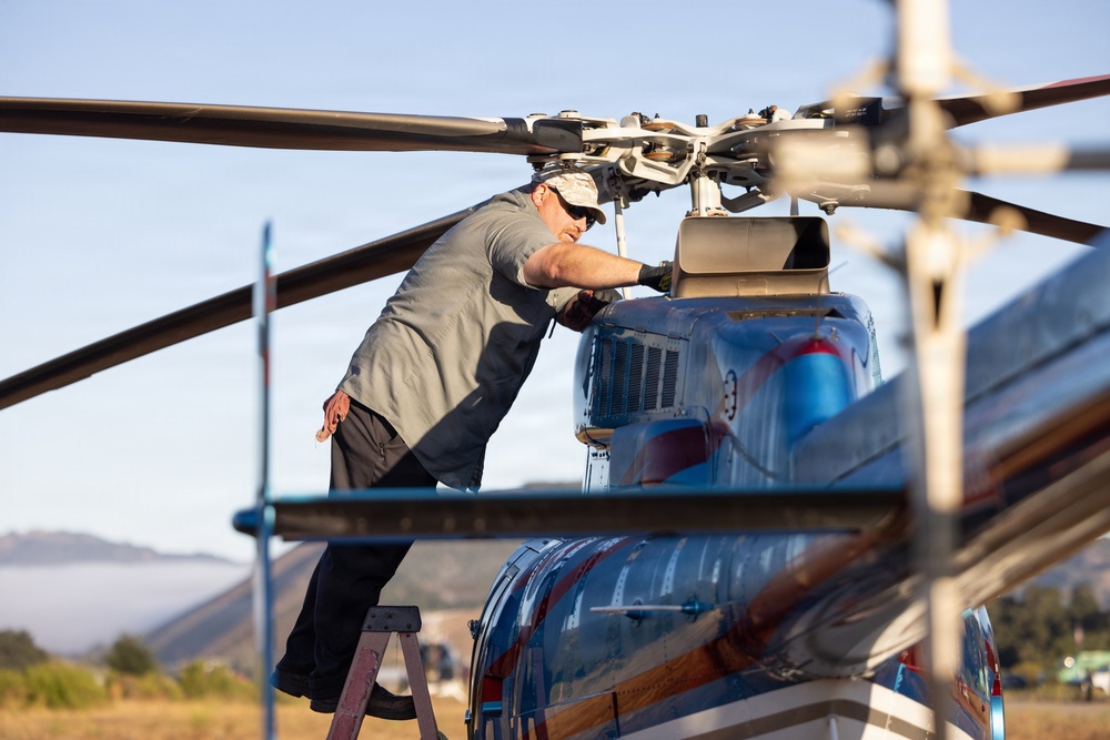 A male technician is performing maintenance on a helicopter, standing on a ladder to access the rotor head assembly at the top of the aircraft. The helicopter has a blue and red metallic finish and appears to be parked on an open airfield with mountains visible in the background. The image is taken during daylight hours with warm, golden lighting suggesting early morning or late afternoon.