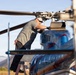 A male technician is performing maintenance on a helicopter, standing on a ladder to access the rotor head assembly at the top of the aircraft. The helicopter has a blue and red metallic finish and appears to be parked on an open airfield with mountains visible in the background. The image is taken during daylight hours with warm, golden lighting suggesting early morning or late afternoon.