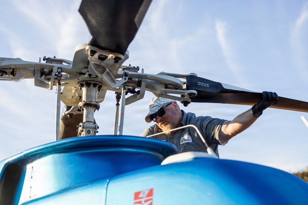 A male technician or mechanic is performing maintenance or a pre-flight inspection on a blue helicopter, gripping one of the main rotor blades. The image is taken from a low angle looking up at the rotor head assembly, providing a detailed view of the mechanical components. The worker is wearing a gray uniform shirt with a logo patch, a camouflage cap, sunglasses, and black gloves.
