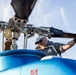 A male technician or mechanic is performing maintenance or a pre-flight inspection on a blue helicopter, gripping one of the main rotor blades. The image is taken from a low angle looking up at the rotor head assembly, providing a detailed view of the mechanical components. The worker is wearing a gray uniform shirt with a logo patch, a camouflage cap, sunglasses, and black gloves.