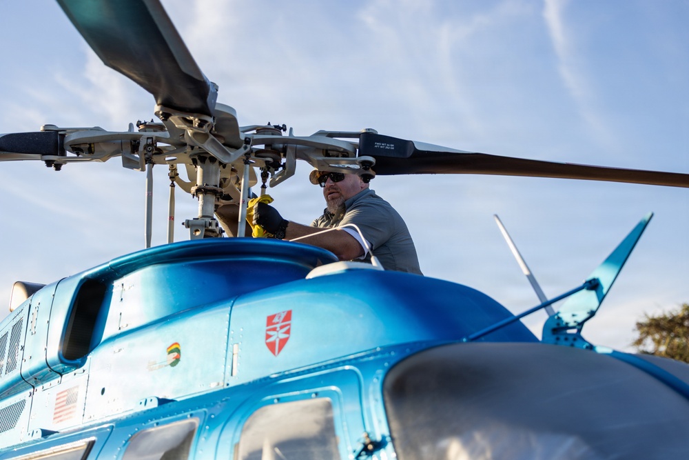 A male technician or mechanic is performing maintenance work on the rotor head assembly of a blue helicopter, viewed from a low angle looking upward. The helicopter features several insignia decals including what appears to be a US Army Corps of Engineers shield emblem and an American flag. The image is taken from ground level, providing a dramatic perspective of both the worker and the rotor hub mechanism.