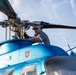 A male technician or mechanic is performing maintenance work on the rotor head assembly of a blue helicopter, viewed from a low angle looking upward. The helicopter features several insignia decals including what appears to be a US Army Corps of Engineers shield emblem and an American flag. The image is taken from ground level, providing a dramatic perspective of both the worker and the rotor hub mechanism.