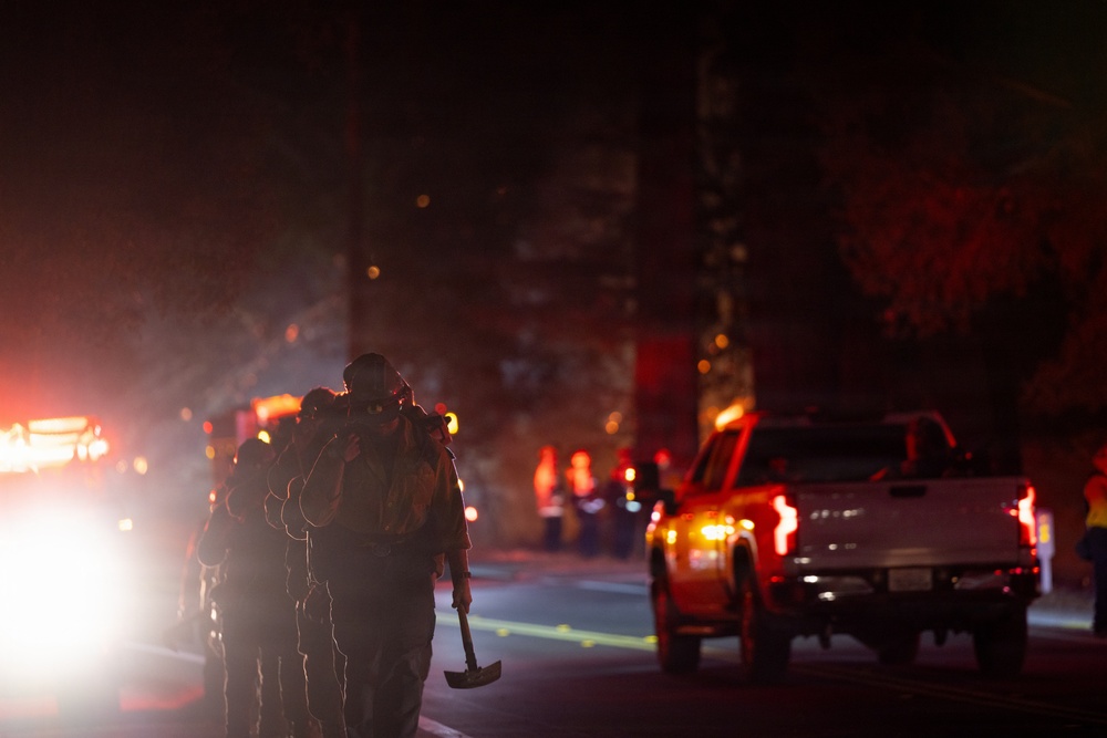 A nighttime emergency response scene showing a group of firefighters or wildland fire crew walking down a road carrying tools, including a visible Pulaski axe/mattock. The scene is illuminated by vehicle headlights, taillights, and what appears to be fire glow in the background. A white pickup truck is visible on the right side of the road amid smoky, hazy conditions.