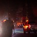 A nighttime emergency response scene showing a group of firefighters or wildland fire crew walking down a road carrying tools, including a visible Pulaski axe/mattock. The scene is illuminated by vehicle headlights, taillights, and what appears to be fire glow in the background. A white pickup truck is visible on the right side of the road amid smoky, hazy conditions.