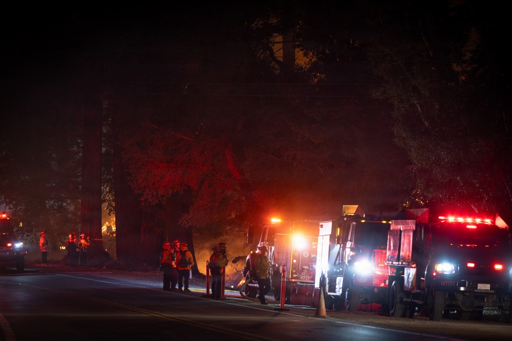 A nighttime emergency response scene showing firefighters and emergency vehicles staged along a road, with visible fire or intense orange glow illuminating trees in the background. Multiple fire trucks and personnel in protective gear are visible on what appears to be a wildfire or structure fire response. The scene is lit primarily by emergency vehicle lights and the ambient glow of the fire.