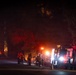 A nighttime emergency response scene showing firefighters and emergency vehicles staged along a road, with visible fire or intense orange glow illuminating trees in the background. Multiple fire trucks and personnel in protective gear are visible on what appears to be a wildfire or structure fire response. The scene is lit primarily by emergency vehicle lights and the ambient glow of the fire.