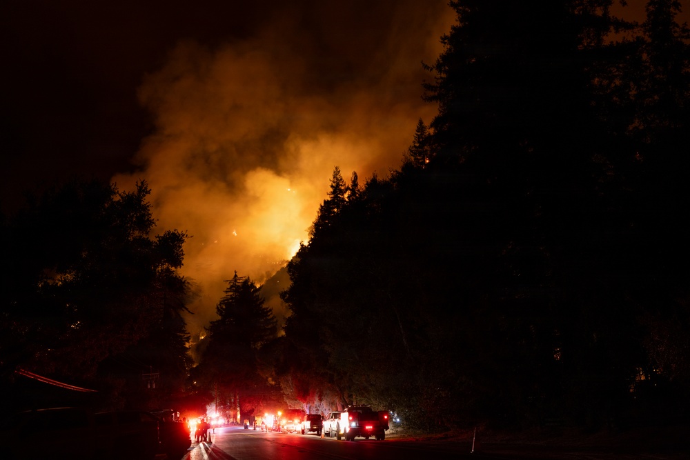 A nighttime wildfire burns intensely on a forested hillside, illuminating the sky with orange and yellow flames and thick smoke. A line of emergency vehicles with red and white lights is staged on a road in the foreground, with personnel visible near the vehicles. The scene depicts an active firefighting response operation at night.