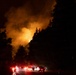 A nighttime wildfire burns intensely on a forested hillside, illuminating the sky with orange and yellow flames and thick smoke. A line of emergency vehicles with red and white lights is staged on a road in the foreground, with personnel visible near the vehicles. The scene depicts an active firefighting response operation at night.