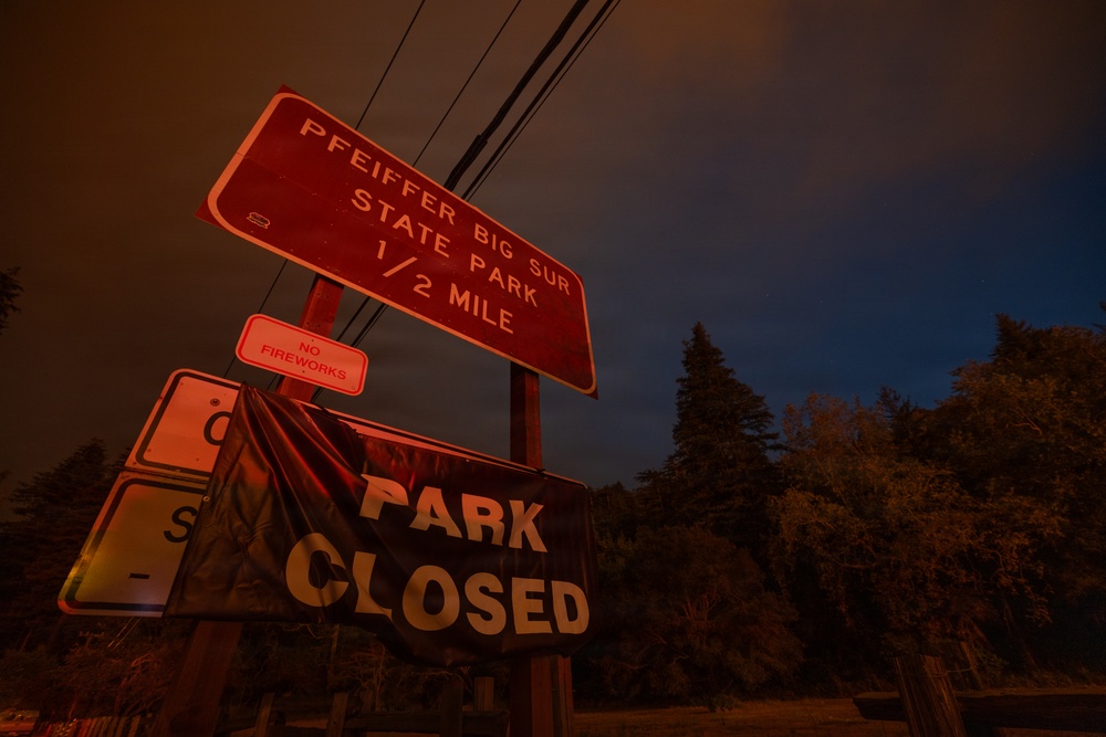 A nighttime photograph showing road signs for Pfeiffer Big Sur State Park, illuminated by an ominous red-orange glow consistent with a nearby wildfire. A 'PARK CLOSED' banner is prominently displayed below the directional sign, along with a 'NO FIREWORKS' warning. The dark sky and reddish atmospheric lighting suggest an active fire emergency in the area.