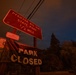 A nighttime photograph showing road signs for Pfeiffer Big Sur State Park, illuminated by an ominous red-orange glow consistent with a nearby wildfire. A 'PARK CLOSED' banner is prominently displayed below the directional sign, along with a 'NO FIREWORKS' warning. The dark sky and reddish atmospheric lighting suggest an active fire emergency in the area.