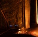A nighttime forest fire photograph captured with a long exposure technique. Ground-level flames and embers are visible along the forest floor, with large tree trunks illuminated by intense orange firelight from the right side. A blue light trail, consistent with a flashlight or torch moved during a long-exposure shot, is visible in the mid-ground.