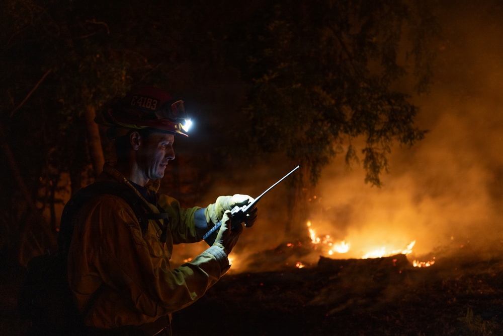 A firefighter is photographed at night while actively monitoring a wildfire. The individual is equipped with a helmet-mounted headlamp and is holding a two-way radio, suggesting coordination with other personnel. Active flames and smoke are visible in the background among trees and brush.