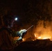 A firefighter is photographed at night while actively monitoring a wildfire. The individual is equipped with a helmet-mounted headlamp and is holding a two-way radio, suggesting coordination with other personnel. Active flames and smoke are visible in the background among trees and brush.
