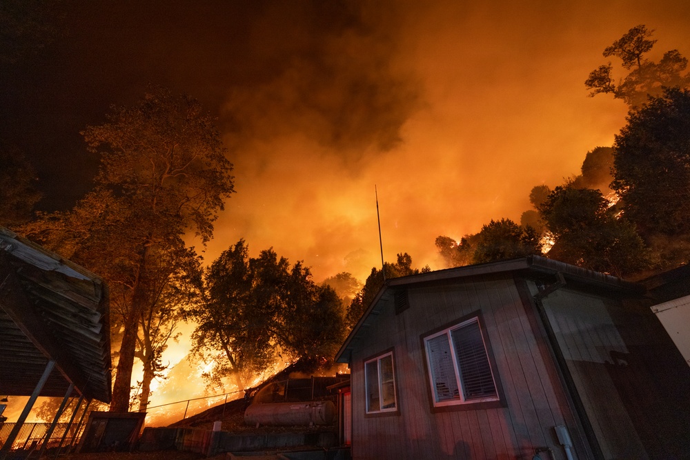 A large wildfire is burning in close proximity to residential structures at night. The sky is illuminated in intense orange and red hues from the flames, which are consuming trees and vegetation on a hillside directly behind the buildings. The structures appear to be in immediate danger as the fire front advances toward them.