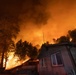 A large wildfire is burning in close proximity to residential structures at night. The sky is illuminated in intense orange and red hues from the flames, which are consuming trees and vegetation on a hillside directly behind the buildings. The structures appear to be in immediate danger as the fire front advances toward them.